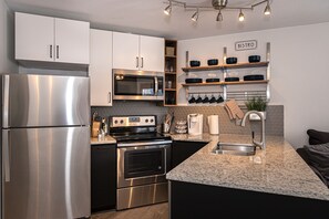 Modern kitchen with stainless steel appliances, white and black cabinetry, open shelving with black dishes, a granite countertop with a sink, and under-cabinet lighting.