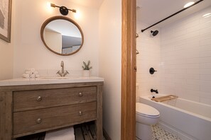 A bathroom featuring a wooden vanity with a round mirror and sink, a potted plant, a white-tiled bathtub with black fixtures, and a folded towel on the sink.