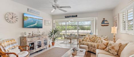 A living room with a coastal theme, featuring a sofa, a wicker chair, a wooden coffee table, a TV, a ceiling fan, and decor. A sliding door opens to a sunroom with a table and chairs.