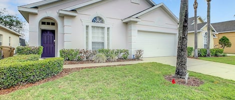 Charming pastel home with a bold purple door, manicured lawn, and spacious driveway.