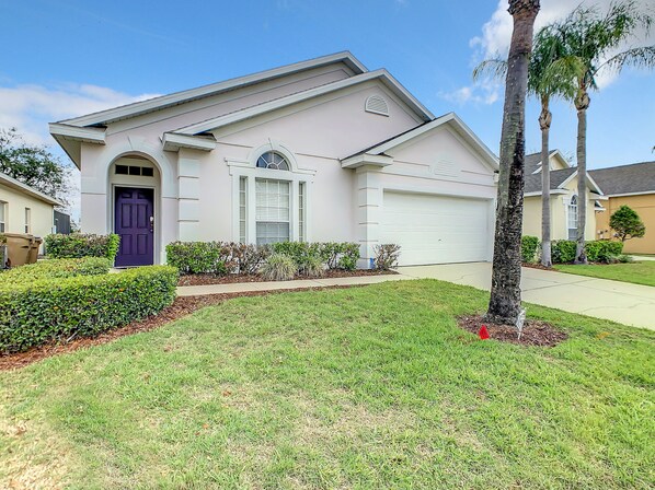 Charming pastel home with a bold purple door, manicured lawn, and spacious driveway.