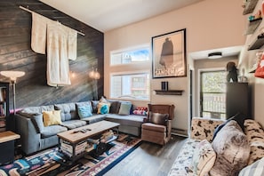 A cozy living room with a large sectional sofa, an armchair, a decorative rug, a wall-mounted TV, shelves, and framed artwork. A shoji screen hangs on a dark wood accent wall.