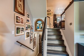 A staircase with dark wood steps and black railings ascends, with artwork and photos on the walls. Upstairs, a well-lit living space is visible.