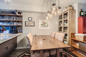 A cozy dining area with a wooden table and cushioned chairs, lit by vintage hanging lights. Shelves stocked with glasses and beverages, a wall clock, and a decorative painting are visible.