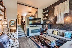 A cozy living room with a large wall-mounted TV, sectional sofa, coffee table, and various decor including shelves of collectibles and a hanging textile art piece. Steps lead to a higher-level area.