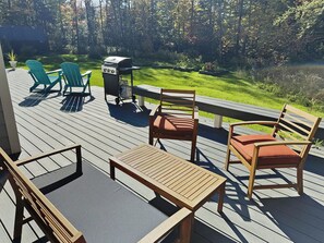 A backyard deck with wooden furniture, two blue lounge chairs, a barbecue grill, and a sunny lawn in the background.