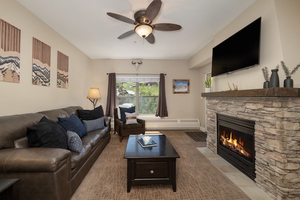 A cozy living room with a leather sofa, coffee table, and stone fireplace. A flat-screen TV is mounted above the fireplace, and artwork decorates the walls. A ceiling fan and lamp provide lighting.