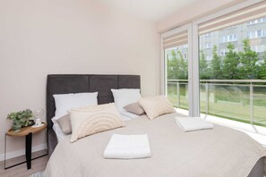 A cozy bedroom with a neatly made double bed, soft bedding in neutral tones, and large windows overlooking greenery.

