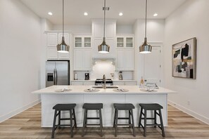 The kitchen area contains an oversized island, 4 barstools, cookware, along with small appliances.