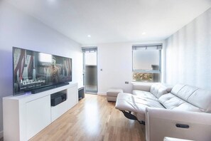 A modern living room on the 12th floor features a 60-inch TV, a dining table with four chairs, and access to a balcony with stunning city views. Two electric reclined sofas provide comfort and a sound system offer entertainment.