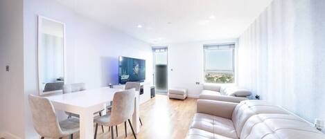 A modern living room on the 12th floor features a 60-inch TV, a dining table with four chairs, and access to a balcony with stunning city views. Two electric reclined sofas provide comfort and a sound system offer entertainment.