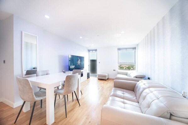 A modern living room on the 12th floor features a 60-inch TV, a dining table with four chairs, and access to a balcony with stunning city views. Two electric reclined sofas provide comfort and a sound system offer entertainment.