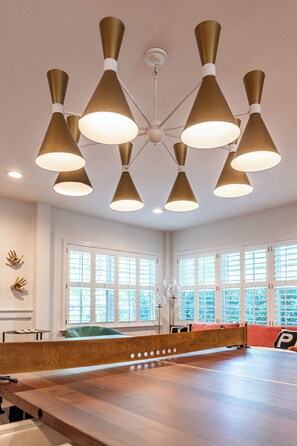 Modern room with a ping pong table and a large, gold chandelier. The room has white walls, large windows with shutters.