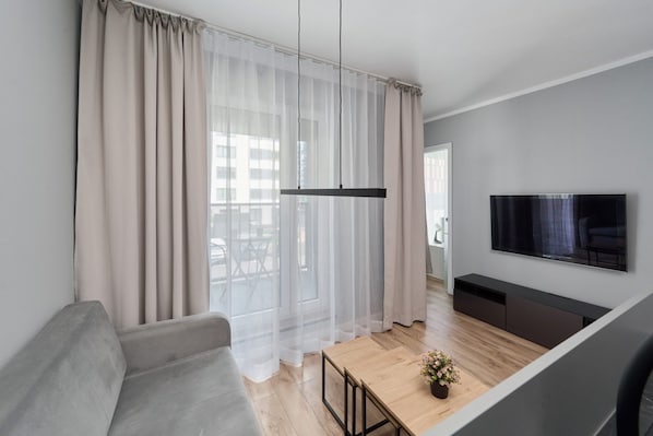 Bright living room with a gray sofa, a TV, and large windows with sheer curtains.