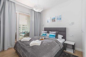 A cozy bedroom featuring a double bed dressed in grey bedding, with curtains and wall art that echo a seaside theme.

