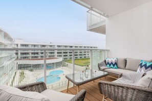 A balcony view of the modern complex and pool area, furnished with a wicker chair and decorative cushions.

