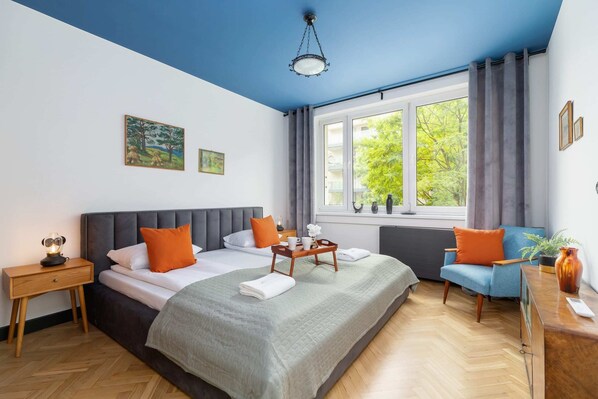 A cozy bedroom with a large bed covered in gray bedding and orange accent pillows. There is a bright window with curtains and a bedside table with a lamp. The wooden floor adds warmth to the room.