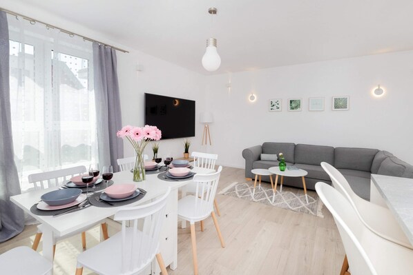 A cozy dining area with a round table set with pink flowers and black plates, placed in a bright, airy room. A television and sofa can be seen in the background.