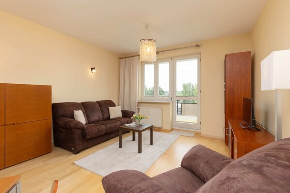 A spacious living room with comfortable brown sofas, a coffee table, and access to a bright balcony.
