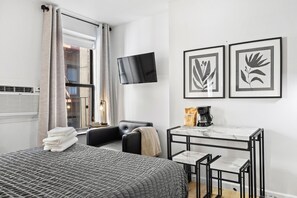 Bed with gray bedding, black chair, table with stools, wall-mounted TV, and framed prints.