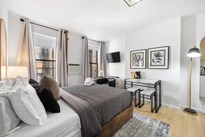 Bright bedroom with white bed, black chair, table with stools, TV, framed prints, AC unit, and curtains.
