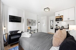 Bedroom with black chair, table with stools, kitchenette, TV, and framed prints. Floor lamp and door visible.