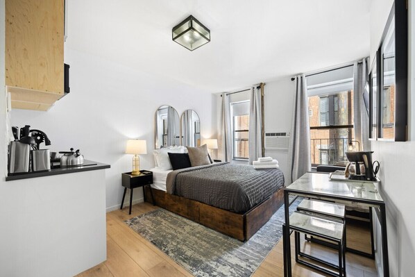Cozy bedroom with a dark wooden bed, gray bedding, nightstands with lamps, a dining table, coffee maker, and white cabinets. Window with curtains and AC unit.
