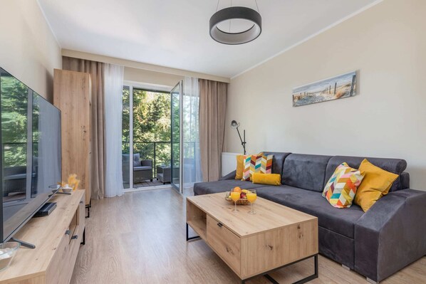 A bright and modern living room featuring a gray sofa with colorful pillows, a wooden coffee table, and a large window with yellow curtains overlooking greenery.