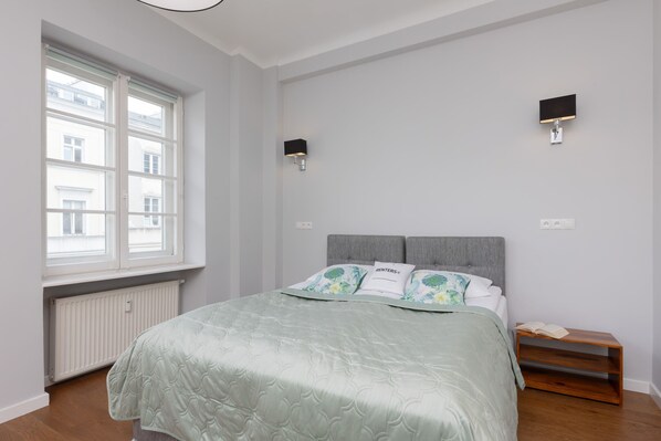 The studio features a comfortable double bed with mint-green bedding, positioned near large windows that fill the room with natural light.