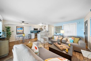 The room is furnished with multiple sofas, each adorned with colorful and tropical-themed pillows. The flooring is a warm, wood-like material that adds a natural element to the space.