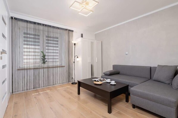 A spacious living room with a gray sofa, a coffee table, and large windows with soft curtains.

