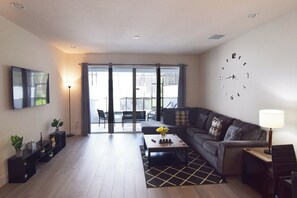 Open-plan living space with L-shaped sofa, large Smart TV, door to the pool deck