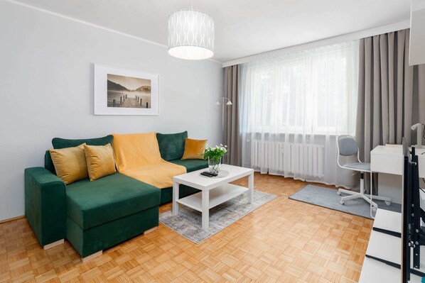 A cozy living room with a green sofa, yellow pillows, and a coffee table, illuminated by natural light.
