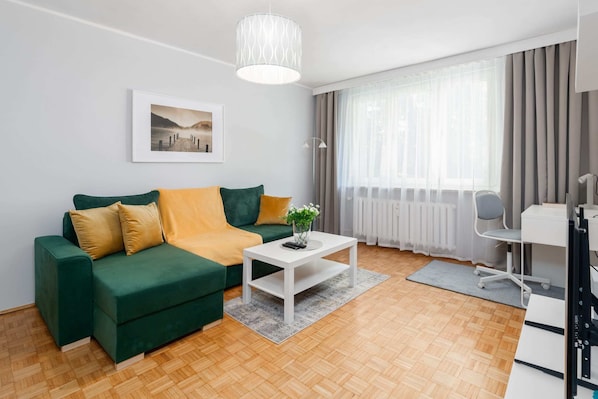 A cozy living room with a green sofa, yellow pillows, and a coffee table, illuminated by natural light.