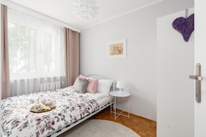 A bright bedroom featuring a bed with floral bedding, a bedside table, and a decorative wall heart.