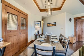 A dining room with a wooden table set with plates and wine glasses. The room features wooden doors, framed art on white walls, and a modern light fixture above the table.