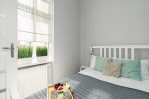 Cozy bedroom with a white bed, pastel pillows, and a simple accent wall, creating a calming atmosphere.

