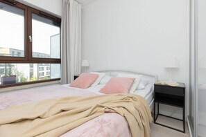 Comfortable bedroom with a soft pink bedspread, large windows, and a modern bedside table.

