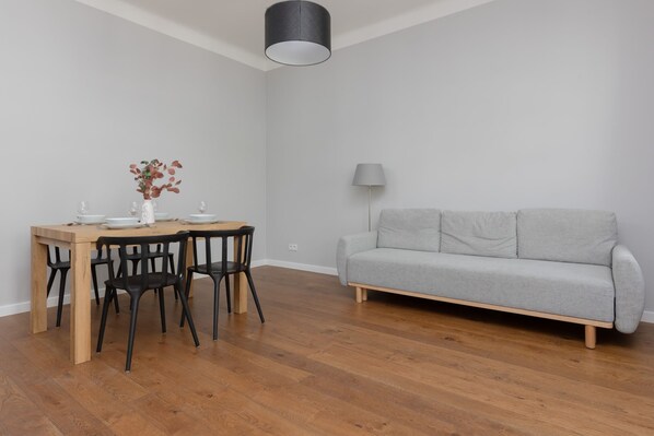A bright and spacious living area with a comfortable gray sofa, wooden flooring, and a dining table with chairs, perfect for relaxing or entertaining.