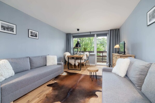 Cozy living room with a soft gray sofa, wooden coffee table, and large windows bringing in natural light.

