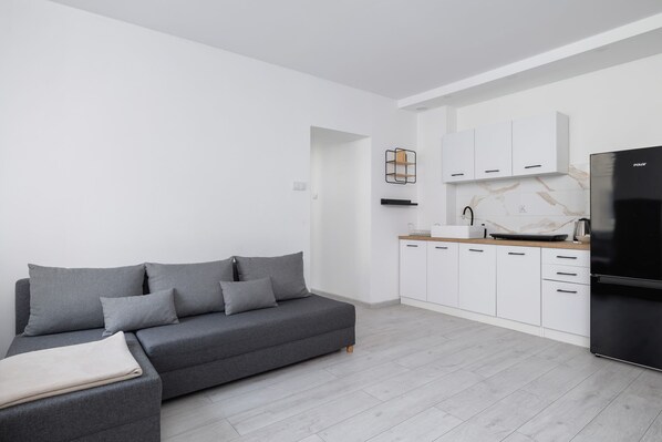 Bright and spacious living area with a grey sofa, open kitchen, and natural light from large windows.

