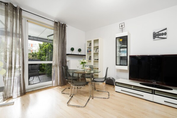 A modern living room with a large TV, glass dining table, and floor-to-ceiling windows providing plenty of natural light.

