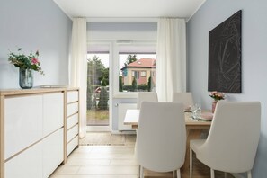A dining area with a white table and chairs, complemented by natural light from large windows.

