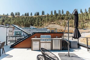 Relax on this rooftop patio with a hot tub, barbecue grill, and wicker chairs under a large umbrella. Enjoy serene views of a tree-covered hillside, with buildings in the distance, all surrounded by a black metal railing for added privacy.
