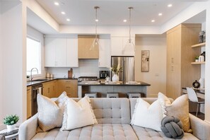This open-concept space features a cozy living room with a beige fabric sofa, white and beige pillows, and a small round table with a potted plant. Perfect for relaxing, the space offers a comfortable and inviting atmosphere for your stay.