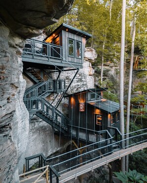 Exterior view of Sandstone Drifter cliff‑house at twilight – sleek all‑black cabin perched against sandstone cliffs, featuring floating staircase and warm outdoor lighting illuminating forest surroundings under a dusky sky.