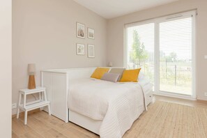 A simple, bright studio featuring a bed with light bedding and a yellow accent pillow, bathed in natural light from the large window.
