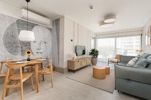 A bright living room with a modern design, featuring a comfortable gray sofa and large windows overlooking greenery.