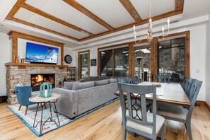 - Cozy open concept living & dining area with large windows framing scenic views
- Stone fireplace, exposed beams, & plush sectional create a warm alpine retreat
- Dining table seats eight under a rustic chandelier, perfect for shared meals & stories