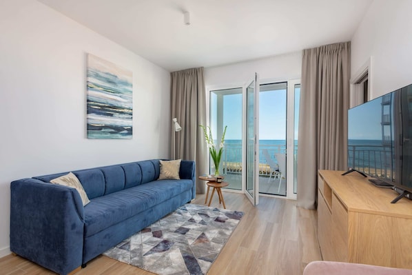 A bright living room with a blue sofa, a glass coffee table, and a balcony with a sea view.